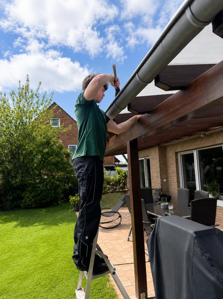 Man on ladder cleaning gutters on residential brick house with patio and garden in background