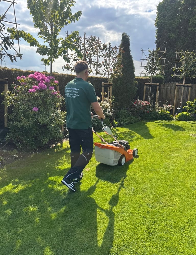 Person in teal shirt mowing a lush green lawn with an orange and white lawn mower in a sunny backyard garden with flowering shrubs and hedges